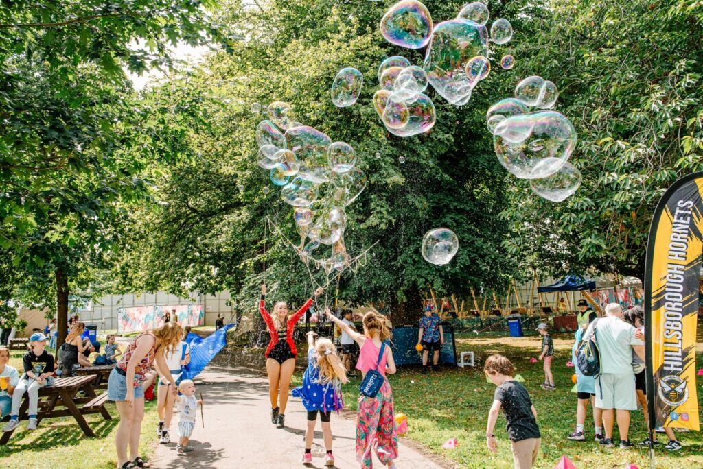 Colorful bubbles float above a lively park where families and performers enjoy a sunny day at Tramlines Festival.