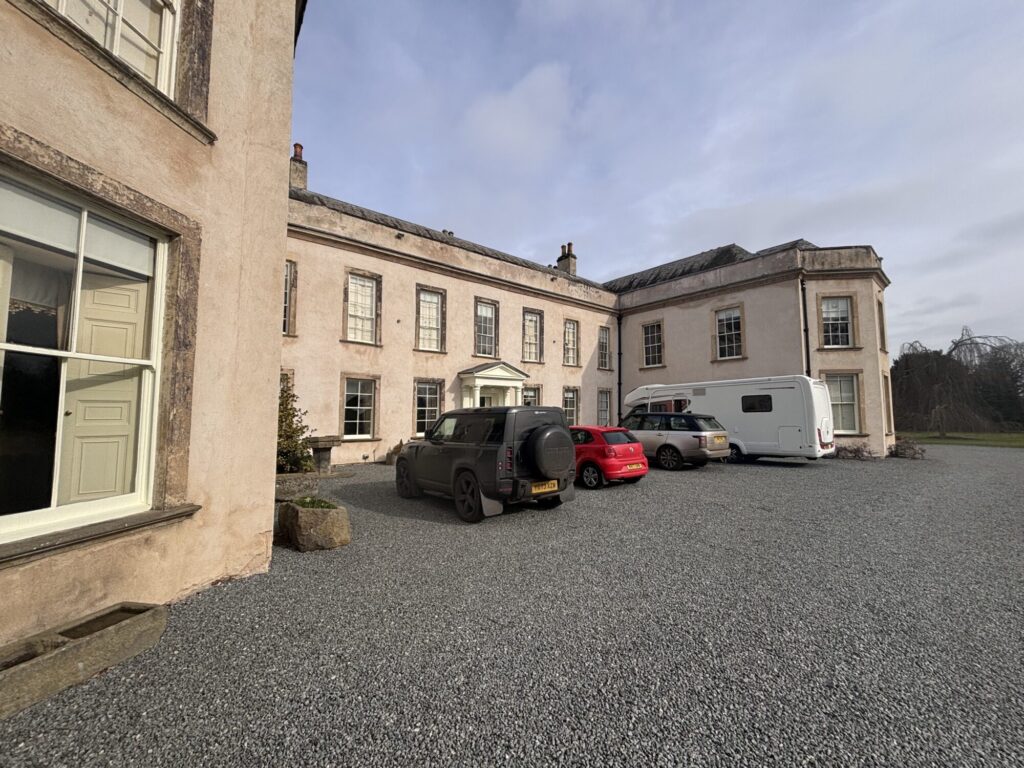 Historic Womersley Park features a pink-stone building with a gravel driveway and parked vehicles under a cloudy sky.