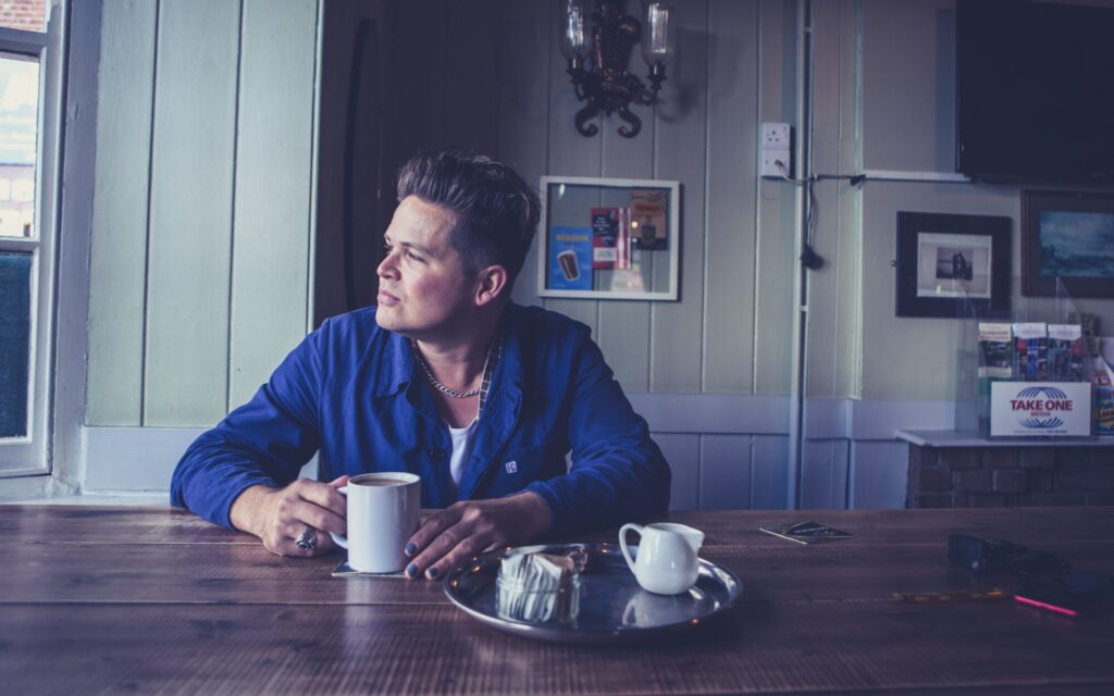 A man in a blue shirt sits at a wooden table, holding a cup, gazing thoughtfully out a window. Soft lighting surrounds him.