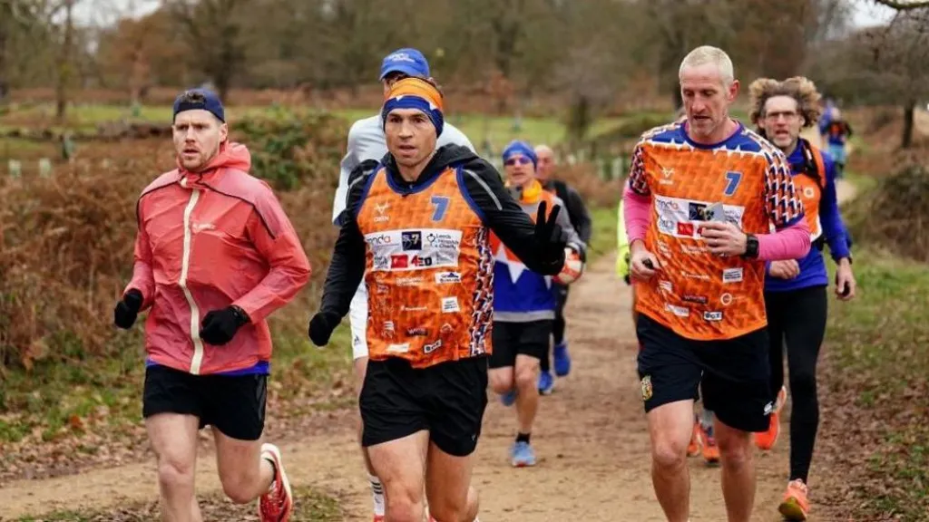 Runners in vibrant orange jerseys, participating in an outdoor challenge along a dirt path surrounded by trees.