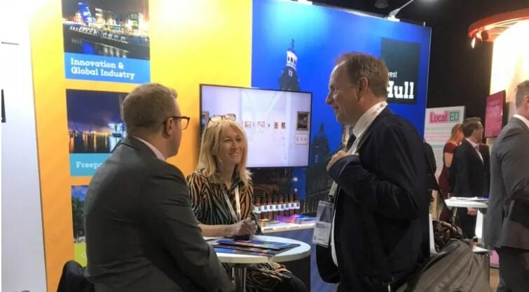 People (two males and a female) talking at a Invest Hull stand in a conference. They look happy and engaged. The males are wearing suits. It looks like a business conference.